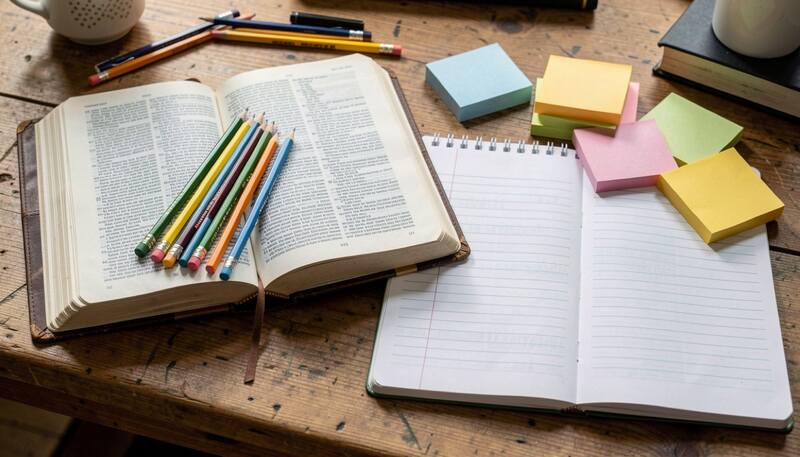 Open Bible with colored pencils, a ruled journal and sticky notes arranged on a wooden desk